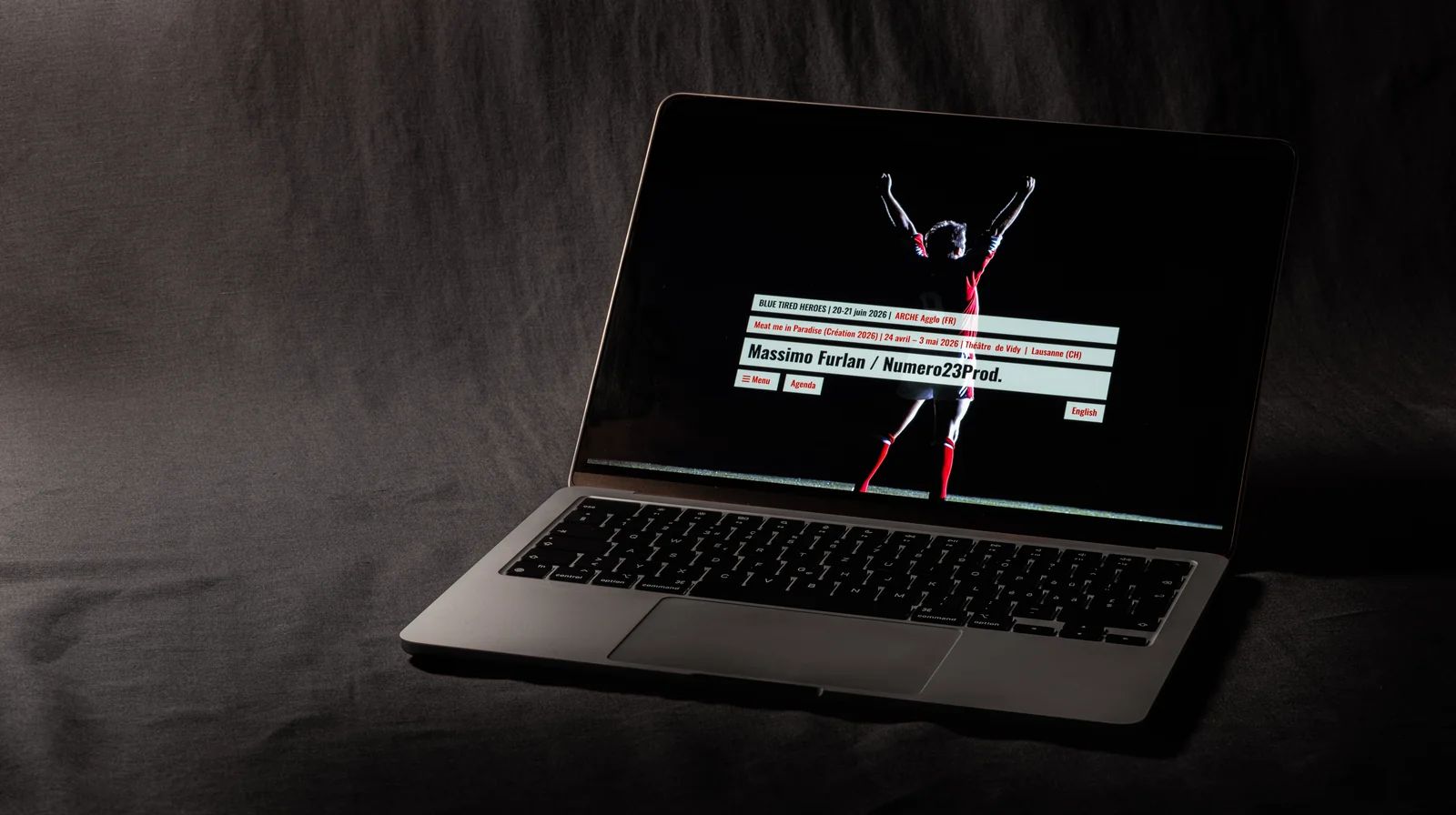 Site web de compagnie pour Massimo Furlan, théâtre et performance : la home de massimofurlan.com sur un MacBook dans la pénombre, une silhouette en maillot rouge bras levés, spectacles et tournées en bandeaux